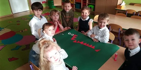 Powiększ grafikę: Dzieci siedzą przy stoliku i wykonują pracę plastyczną pt. Smok Wawelski. Do zielonego brystolu przyklejają czerwone oraz białe trójkąty. Mają przedstawiać kolce oraz zęby smoka.