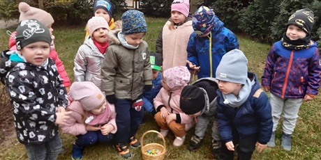 Powiększ grafikę: Dzieci znajdują się w ogrodzie przedszkolnym. Poszukują czekoladowych zajek ukrytych przez wielkanocnego zajączka.  Dzieci stoją nad koszykiem, w którym znajdują się znalezione czekoladowe jajeczka.