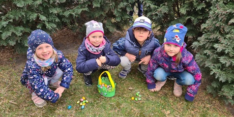 Powiększ grafikę: Dzieci znajdują się w ogrodzie przedszkolnym. Poszukują czekoladowych zajek ukrytych przez wielkanocnego zajączka. Czworo dzieci kuca nad koszyczkiem z czekoladowymi pisankami. Kilka pisanek leży prze
