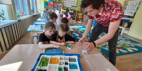 Powiększ grafikę: Dzieci znajdują się w sali przedszkolnej. Na pierwszym planie przy stole siedzą dwie dziewczynki. Nad nimi po prawej stronie zdjęcia stoi instruktor. Na stole znajduje się pudełko z klockami lego. Dzi