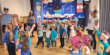 Powiększ grafikę: Na sali gimnastycznej odbywa się Morski bal. Dzieci mają założone na głowach opaski i ubrane są na niebiesko. W tle znajduje się dekoracja przedstawiająca morski krajobraz.