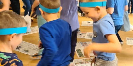 Powiększ grafikę: Na sali gimnastycznej odbywa się Morski bal. Dzieci mają założone na głowach opaski i ubrane są na niebiesko. Dzieci biorą udział w konkursie, i ich zadaniem jest stanąć na gazecie rozłożonej na podło