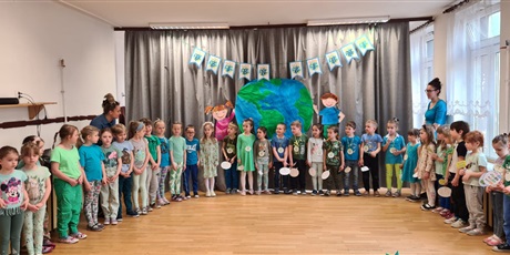Powiększ grafikę: Dzieci znajdują się na sali gimnastycznej, stoją w półkolu. Nad nimi znajduje się napis Dzień Ziemi oraz dekoracje przedstawiające kulę ziemską i trzymające ją dzieci.