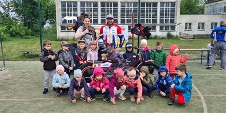 Powiększ grafikę: Dzieci pozują do pamiątkowego zdjęcia z motocyklem żużlowym. Na zdjęciu oprócz dzieci znajduje się menadżer klubu GKS Wybrzeże Gdańsk oraz wychowawca. Wszyscy znajdują się na szkolnym boisku.