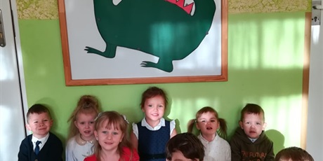 Powiększ grafikę: Dzieci siedzą pod tablicą, na której zawieszony jest Smok Wawelski. Dzieci wykonały go samodzielnie z zielonego i czerwonego brystolu