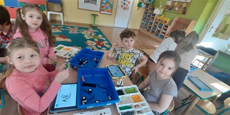 Powiększ grafikę: Dzieci znajdują się w sali przedszkolnej. Siedzą przy stoliku, na którym leżą pojemniki z klockami Lego. Troje dziewczynek oraz jeden chłopiec kontrują wiatraki. Odbywają się zajęcia z robotyki.
